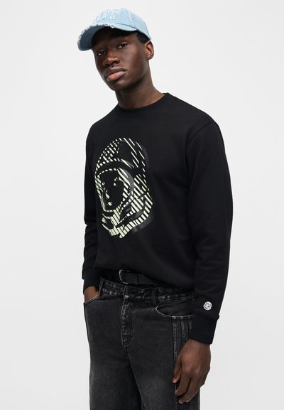 Μαύρο φούτερ με μεγάλο γραφικό αστροναύτη, συνδυασμένο με γκρι τζιν παντελόνι. Ένα ανοιχτό μπλε καπέλο με φθορές ολοκληρώνει την εμφάνιση.