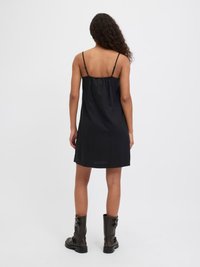 Vestido preto sem mangas com alças ajustáveis, apresentando um corte solto e uma textura suave, combinado com botas pretas de fivela até ao joelho.