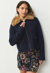 Cappotto corto blu navy con colletto in pelliccia ecologica, dotato di cerniera frontale e due tasche a toppa. La texture è morbida, con un design pulito e su misura.
