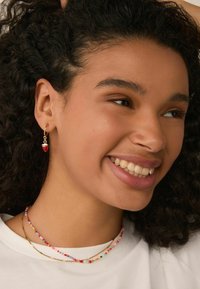 Gold hoop earrings with a red strawberry charm, complemented by colorful beaded necklaces on a model wearing a plain white shirt.