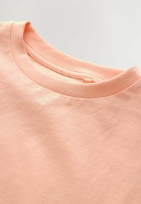 Pfirsichfarbiges Baumwoll-T-Shirt mit rundem Ausschnitt, glatter Textur und sauberem Nahtdetails am Kragen. Keine sichtbaren Grafiken oder Muster.