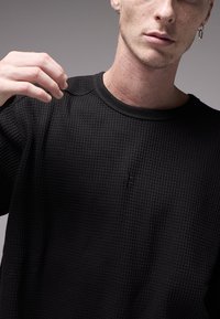 Camisola preta de malha em padrão waffle com decote redondo, detalhe discreto do logótipo no peito e corte descontraído. Tecido texturizado e leve.