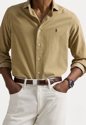 Mężczyzna ubrany w beżową koszulę na guziki z podwiniętymi rękawami, białe spodnie, brązowy skórzany pasek i niebieski zegarek na rękę, ręce w kieszeniach.