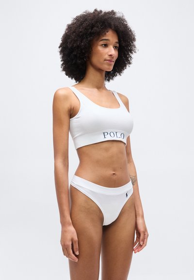 Γυναίκα με ίσια μαλλιά που φοράει λευκό αθλητικό σουτιέν με την επιγραφή "POLO" και αντίστοιχα λευκά σλιπ, στέκεται μπροστά σε απλό φωτεινό φόντο.