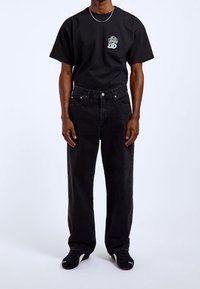 Czarny bawełniany T-shirt z małą wielokolorową grafiką lwa, zestawiony z luźnymi czarnymi jeansami i czarnymi trampkami.