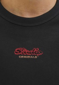 Čierne bavlnené tričko s rebrovaným okrúhlym výstrihom. Na hrudi má viacfarebnú nálepku "Studio" v červenej a "ORIGINALS" v zlatej farbe.