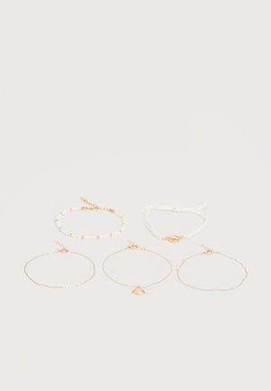 Cinq bracelets délicats en or et blanc avec de petites perles et des charms en coquillage, disposés sur une surface claire en deux rangées.