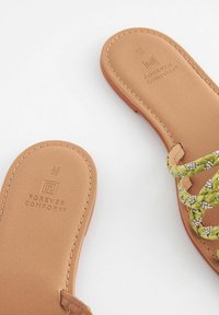 Platta sandaler i tan-färg med flätade gröna detaljer och små dekorativa element. Har en mjuk och texturerad fotbädd med varumärke på sulorna.