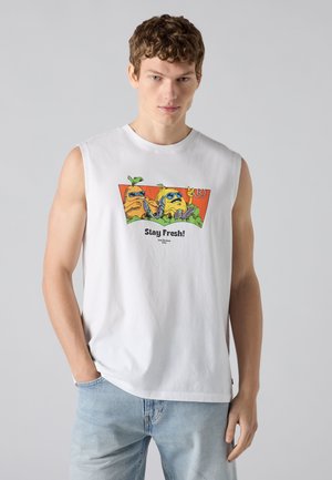 Jeune homme portant un débardeur blanc avec des fruits dessinés en cartoon avec des lunettes de soleil et le texte « Stay Fresh ! » rentré dans un jean bleu clair.
