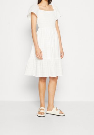 Woman wearing a white knee-length dress with puffed short sleeves and white sandals with buckles, standing against a plain white background.