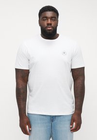 Weißes Baumwoll-T-Shirt mit kurzen Ärmeln und rundem Halsausschnitt, das auf der linken Brust ein kleines grafisches Motiv eines Auges mit Sonnenstrahlen zeigt. Kombiniert mit hellblauen Jeans.