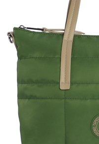 Bolsa de nylon verde con textura acolchada, asas de cuero beige y un parche con logo circular en el frente. Cierre con cremallera en la parte superior.