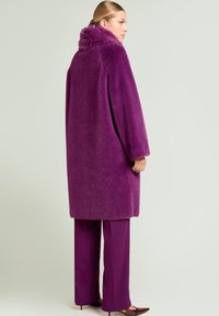 Abrigo de piel sintética púrpura con cuello alto, mangas anchas y corte amplio, combinado con pantalones púrpuras a juego y zapatos de punta fina.