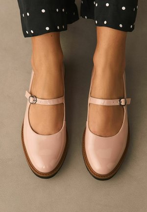 Pieds portant des chaussures Mary Jane en cuir verni rose pâle avec des brides à boucle, associées à un pantalon noir à pois blancs.