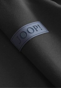 Tela negra con una textura suave que presenta un parche rectangular azul con el logotipo en relieve de la palabra "JOOP!" en letras mayúsculas.