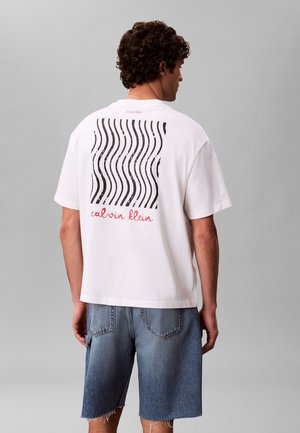 Muž nosí biele tričko s čiernymi vlnitými líniami a červeným textom "calvin klein" na chrbte, v kombinácii s modrými denimovými šortkami, stojí na sivom pozadí.