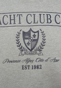 Λογότυπο Yacht Club Monaco με ασπίδα, κορώνα, fleur-de-lis, κλαδιά δάφνης και κείμενο "Provence Alpes Côte d'Azur EST 1982" σε γκρι ύφασμα.