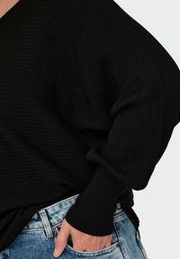 En svart, ribbad tröja hänger över en hand som vilar i fickan på blekta blå jeans, vilket framhäver en avslappnad och stilig klädstil.