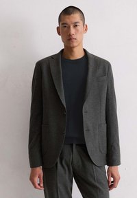 Man in een donkergrijs pak met bijpassende broek en een zwarte ronde hals t-shirt, leunend tegen een witte muur.