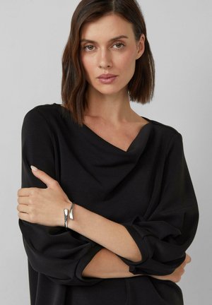 Vrouw met kort bruin haar, gekleed in een zwarte top en een moderne zilveren armband, met gekruiste armen, neutrale grijze achtergrond.