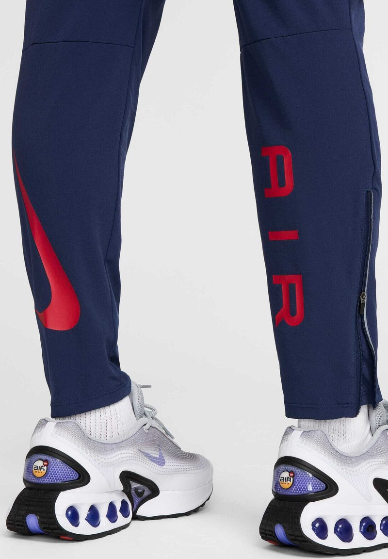 Granatowe spodnie sportowe z czerwonym napisem "AIR" i dużym czerwonym logo Nike. Połączone z białymi i szarymi sneakersami z purpurowymi akcentami.