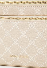 Beige texturerad handväska med vita cirkulära mönster, gyllene dragkedjor och en logotyp präglad i guld på framsidan. Kompakt design som passar för det mest nödvändiga.