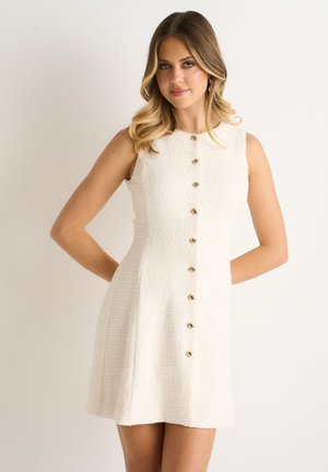 Woman with wavy blonde hair wears sleeveless textured white dress with gold buttons, standing against a plain light background.