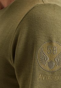 Majica odmaslinasto zelene boje s pletenom teksturom, okruglim izrezom i kružnom oznakom s "58" i krilima na lijevom ramenu.