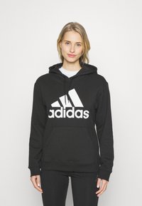 Hoodie noir en tissu doux, doté d'une poche kangourou, d'une capuche à cordon de serrage et d'un grand logo Adidas blanc sur la poitrine.