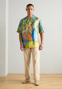 Camisa de botones de manga corta con un vibrante patrón abstracto en azul y amarillo. Combinada con pantalones beige y zapatos slip-on claros.