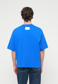 Μπλε βαμβακερό μπλουζάκι με κοντά μανίκια, στρογγυλή λαιμόκοψη και ορθογώνια λευκή ετικέτα στην πλάτη. Απλό σχέδιο, χωρίς μοτίβα ή διακοσμητικά στοιχεία.