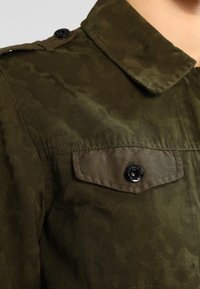 Camicia militare verde oliva con un motivo di camouflage testurizzato, dotata di una tasca sul petto con bottone e dettagli in bottone neri.