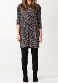 Nainen, jolla on päällään musta-valkoinen paisley-kuosisia pitkä tunika, mustat kiinteät housut ja mustat nilkkurit valkoista taustaa vasten.