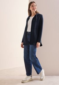 Navy hooded jacket with a zip front, featuring two side pockets. Worn with white t-shirt and blue denim jeans, paired with white sneakers.