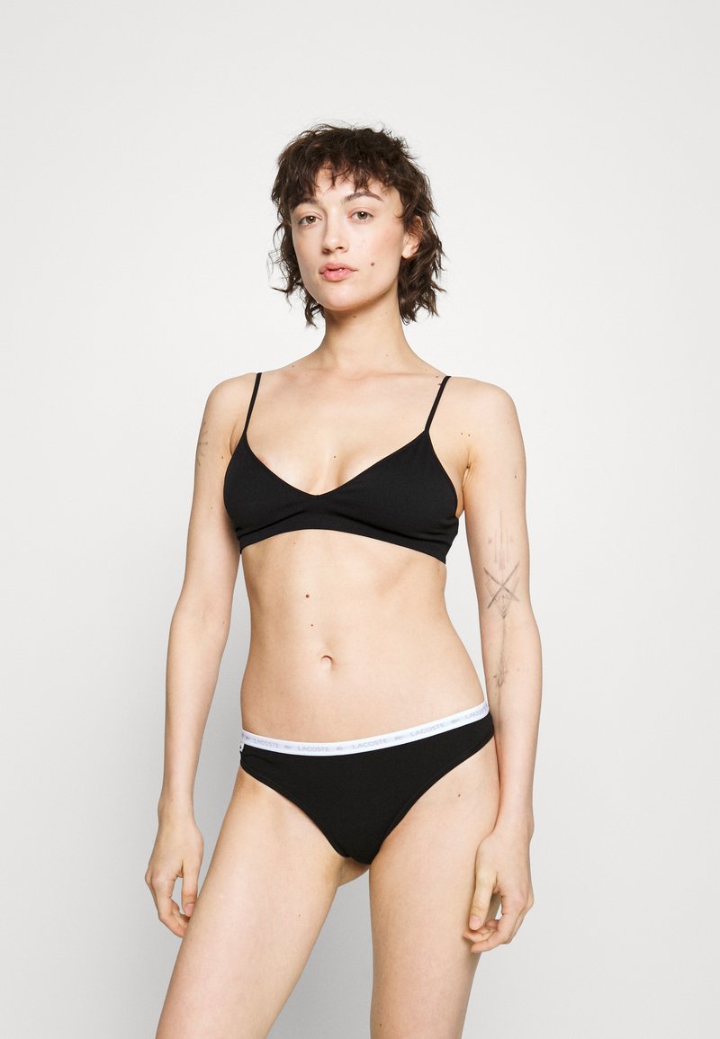 Brassière noire avec des bretelles fines ajustables et un design en V, associée à un slip noir assorti avec une ceinture de marque blanche.