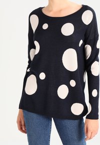 Navy sweater featuring large cream polka dots, round neckline, soft texture, and relaxed fit. Paired with denim jeans.