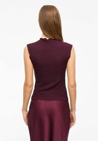 Bordeaux ärmlös topp med texturerad tyg och volangkrage, i kombination med en slät bordeauxkjol som framhäver en figurnära silhuett.