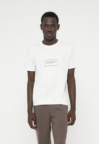 Biele tričko s krátkym rukávom s obdĺžnikovým čiernym logom v strede. Mäkká tkanina, klasický okrúhly výstrih a voľný strih.