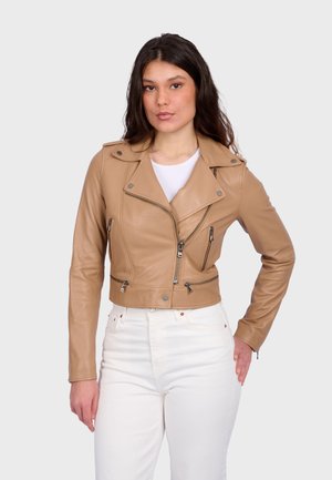 Jonge vrouw die een bruine leren motorjack draagt over een wit shirt en witte, hooggetailleerde broek, staand met één hand op haar heup.