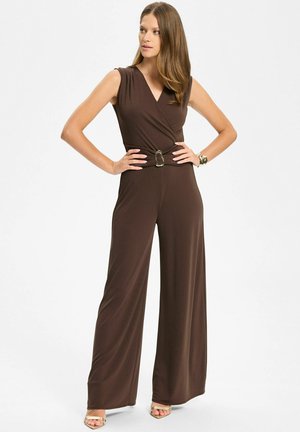 Frau posiert in einem ärmellosen braunen Jumpsuit mit V-Ausschnitt, weitem Bein, Taillengürtel, goldenem Armband und offenen goldenen Absätzen vor weißem Hintergrund.