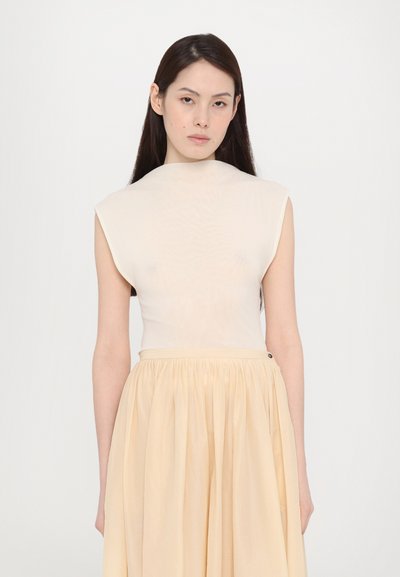 Beige, ujj nélküli felső, magas gallérral, sima anyagból készült. Egy világos sárga, laza, redőzött szoknyával párosítva. Egyszerű és minimalista design.