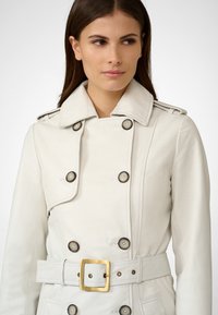 White leather trench coat with double-breasted buttons, structured collar, and a belt featuring a square gold buckle. Smooth texture and close fit.