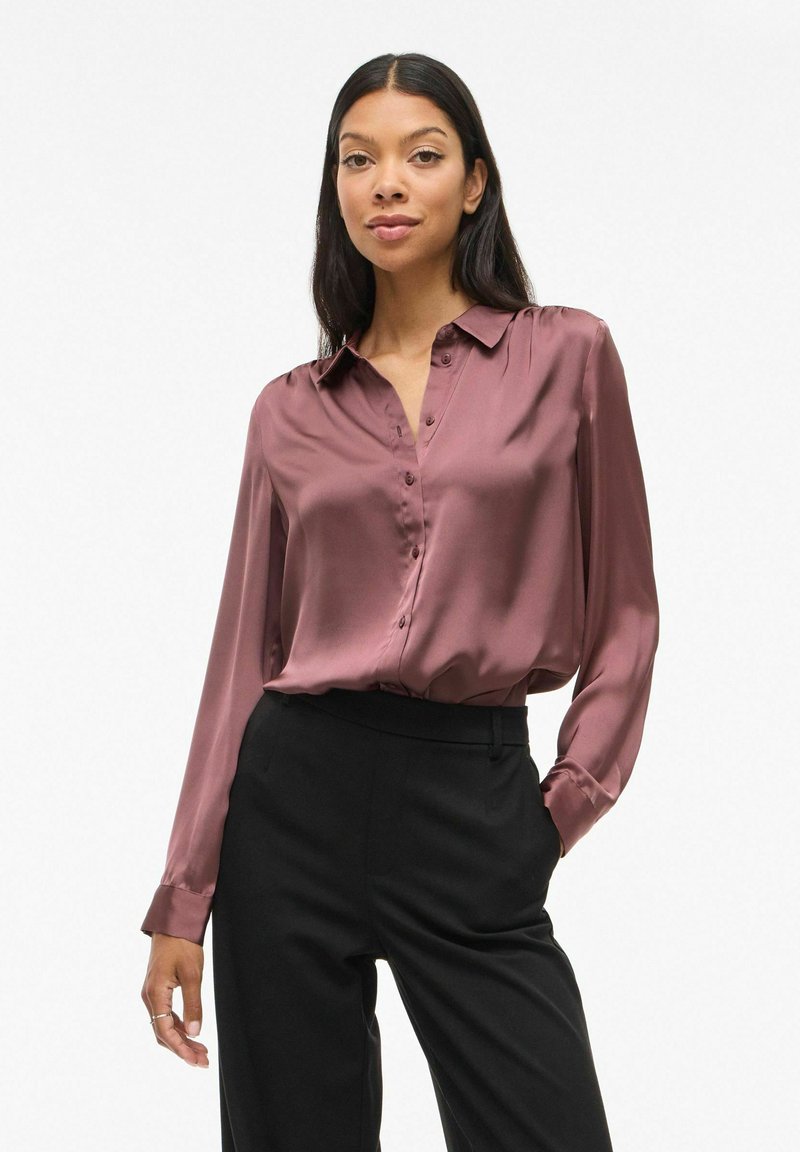 Blusa in satin bordeaux con colletto, maniche lunghe e chiusura a bottoni. Indossata con pantaloni neri a vita alta. Texture liscia e vestibilità sartoriale.