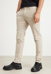 Béžové kalhoty na míru s hladkou texturou, úzký střih, postranní kapsy, ohrnuté lemy, v kombinaci s černými společenskými botami s robustní podrážkou.