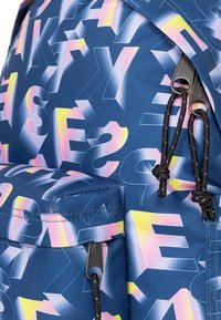 Mochila con una tela azul que presenta letras geométricas grandes y multicolores. Los detalles notables incluyen tiradores de cremallera negros y una superficie texturizada.