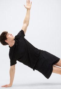 Μαύρο αθλητικό πουκάμισο και σορτς σε άτομο σε θέση πλάγιας σανίδας, με ένα χέρι σηκωμένο. Λείο ύφασμα, χαλαρή εφαρμογή, μίνιμαλ σχεδιασμός.