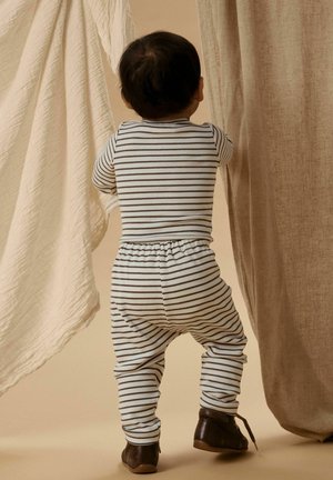 Bambino piccolo con pigiama a righe e scarpe marroni in piedi che tiene tende beige e crema all'interno.