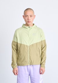 Lichte, bicolor jas in lichtgroen en kaki, met een capuchon, een ritssluiting aan de voorkant en een logo op de borst, met een gladde textuur.