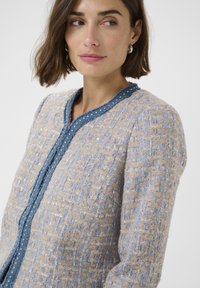 Beige and blue textured jacket with a woven pattern, featuring a blue trim and small decorative beads along the front.