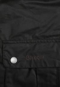 Zwarte jas met een voorkeurszak met een zilveren drukknoop en geborduurd "Barbour" logo in donkere draad op de stof. Zachte textuur.